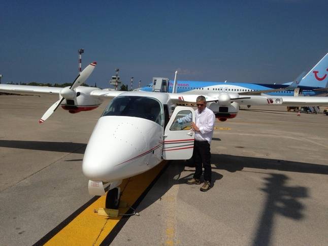 2009 tecnam p2006 parked on airifeld with large aircraft in view man standing nexto plane with cockpit door opne white body work with red and grey stripes